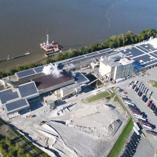 A manufacturing facility sits by a river. Across the roof are solar panels. 