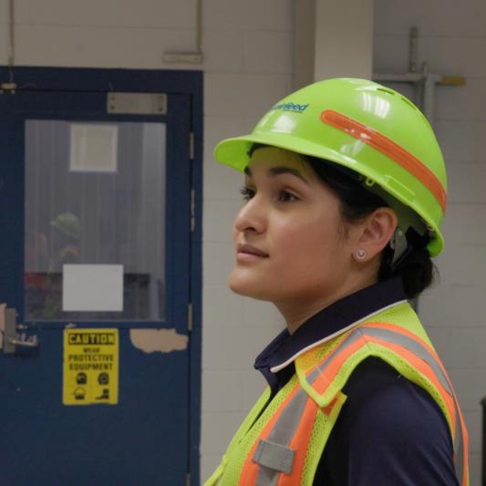 A woman wearing a yellow hard hat and safety vest stands in the manufacturing plant