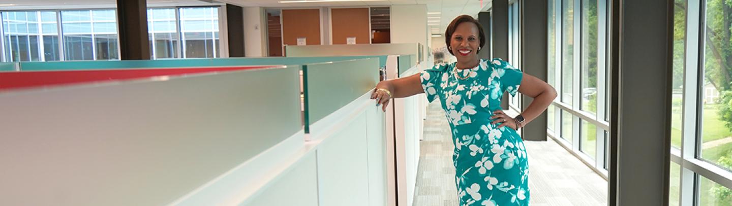 A Black woman wearing a green dress stands near cubicle walls in an office environment