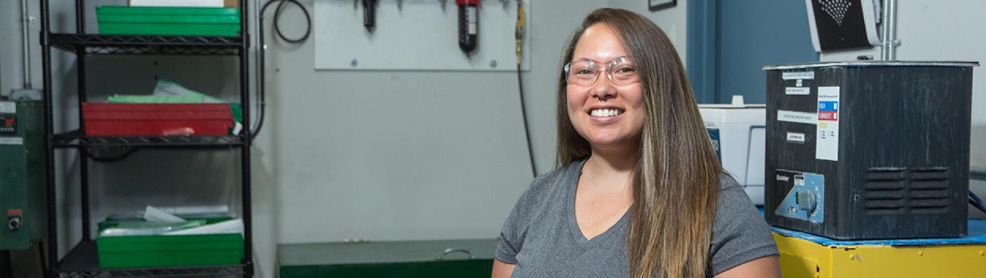 An Asian woman stands in a manufacturing plant setting, wearing safety goggles