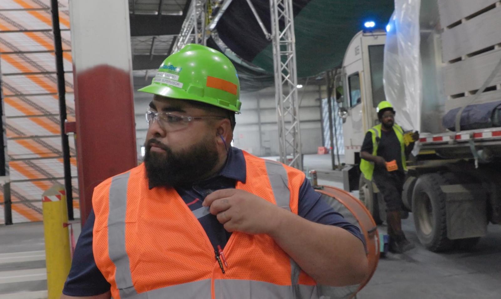 A man with a yellow hard hat and orange vest is on manufacturing plant floor